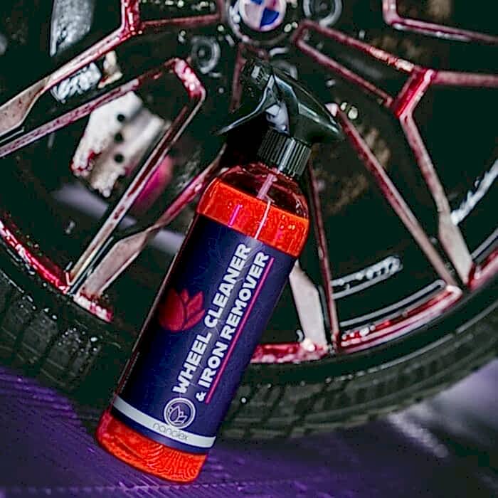 Une bouteille de nettoyant pour jantes et décapant pour fer Nanolex repose sur le sol près dune roue de voiture, en partie recouverte dun liquide rouge. Son étiquette présente un design violet accrocheur avec un texte blanc clair et rouge vif. - Tekalab
