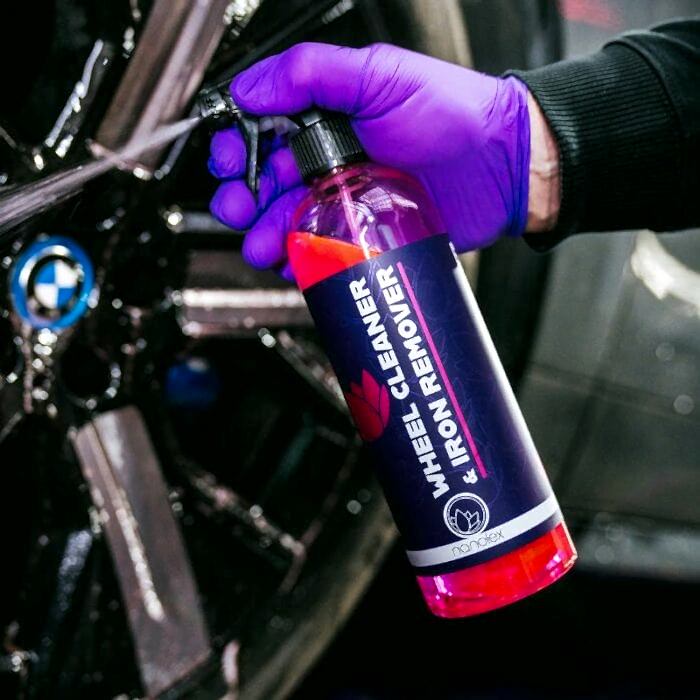 Une personne portant des gants violets tient un vaporisateur Nanolex Wheel Cleaner & Iron Remover avec un nettoyant rose vif devant une roue de voiture noire, avec un logo bleu et blanc. - Tekalab
