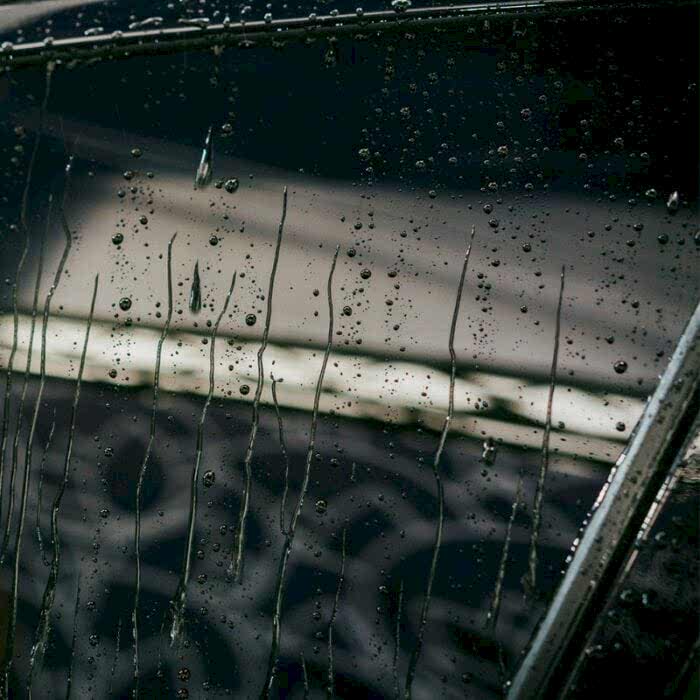 Les gouttes de pluie coulent sur une surface en verre, tout comme le spray Nanolex Si3D (200 ml) crée des motifs protecteurs avec sa technologie céramique, réfléchissant la lumière sur les gouttelettes et mettant en valeur leur brillance texturée sur un fond sombre et légèrement flou. - Tekalab